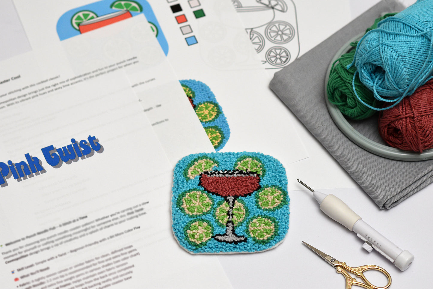 Embroidery hoop, a punch needle coaster with a cocktail design, yarn, and sewing tools on a table.