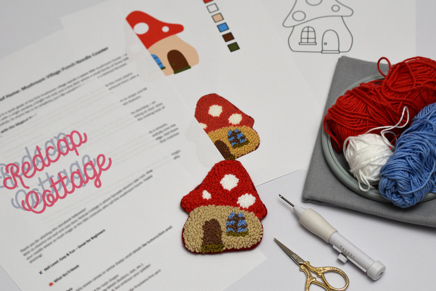 Embroidered punch needle coaster with mushroom shaped house design on paper with yarn and sewing tools.