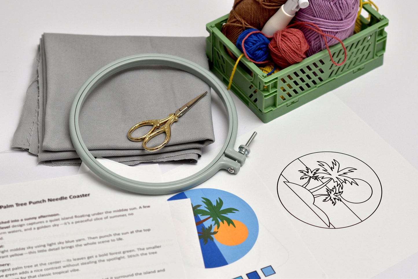 Embroidery hoop, scissors, fabric, yarn, and a small crate on a white surface with a palm tree design.