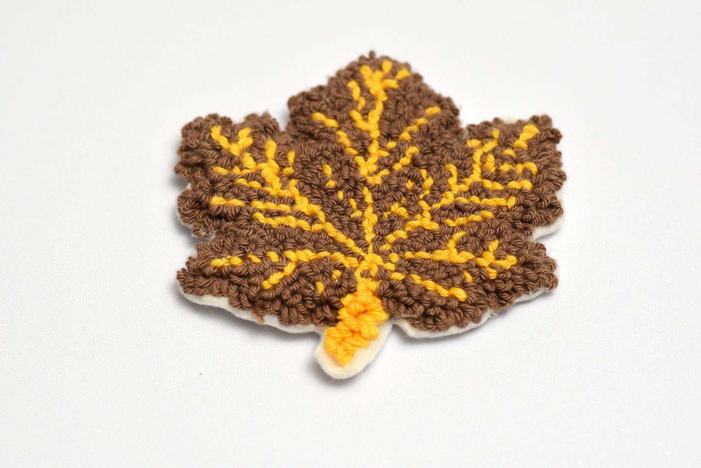 Punch needled leaf-shaped coaster with brown and yellow colors on a white background
