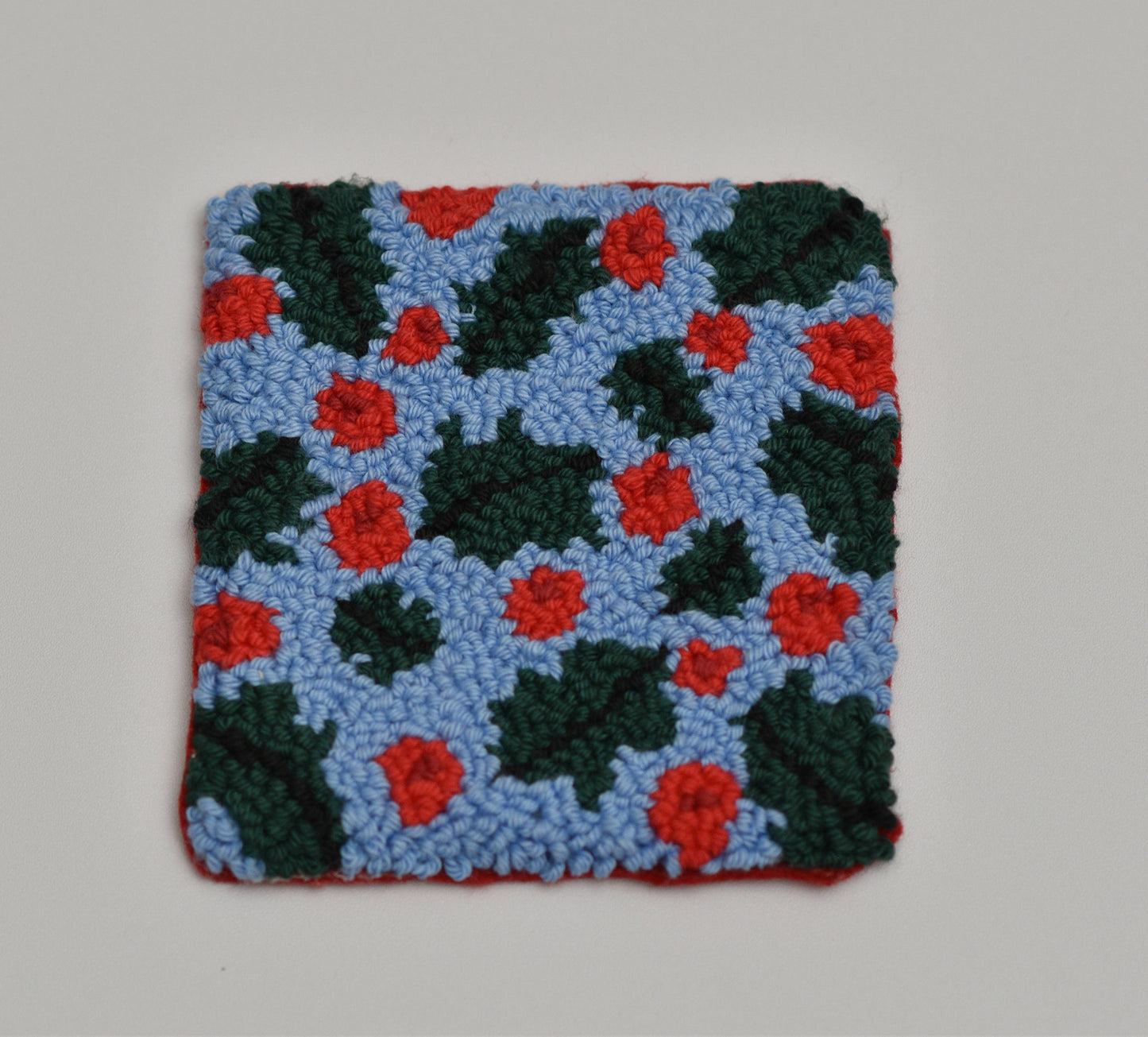 Square punch needle coaster with mistletoe pattern featuring red flowers on a blue background